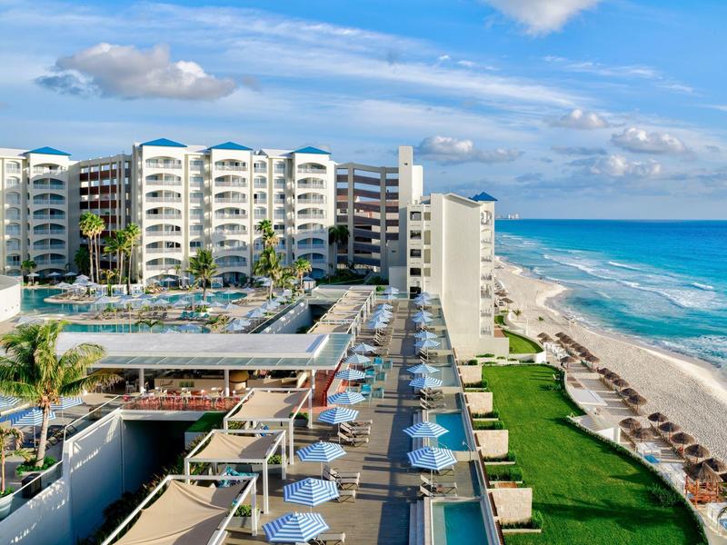 Mehrstöckiges Hotel am Strand mit Pools, Sonnenschirmen und blauem Ozean im Hintergrund.