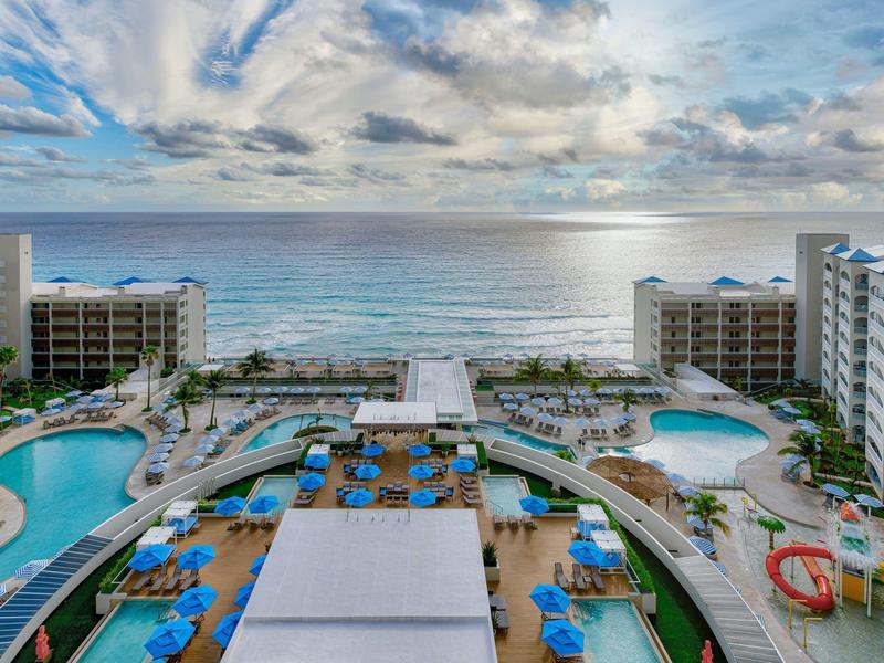 Luxuriöses Resort mit Pools, Liegestühlen und Blick auf das blaue Meer unter wolkigem Himmel.