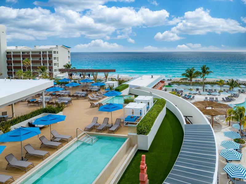 Strandhotel mit Pool, blauen Sonnenschirmen und Liegestühlen vor türkisfarbenem Meer und blauem Himmel.