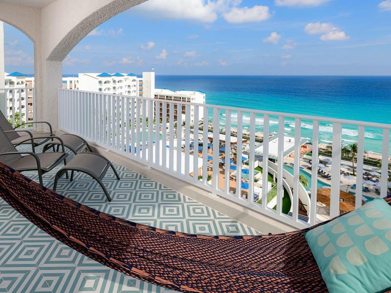 Balkon mit Hängematte, zwei Stühlen und Meerblick unter blauem Himmel und weißen Wolken.
