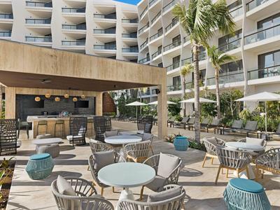 Moderne Hotelterrasse mit grauen Stühlen, runden Tischen, blauen Hockern und Palmen.