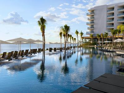 Luxusní hotelový bazén s lehátky, palmami a výhledem na oceán za jasného nebe.