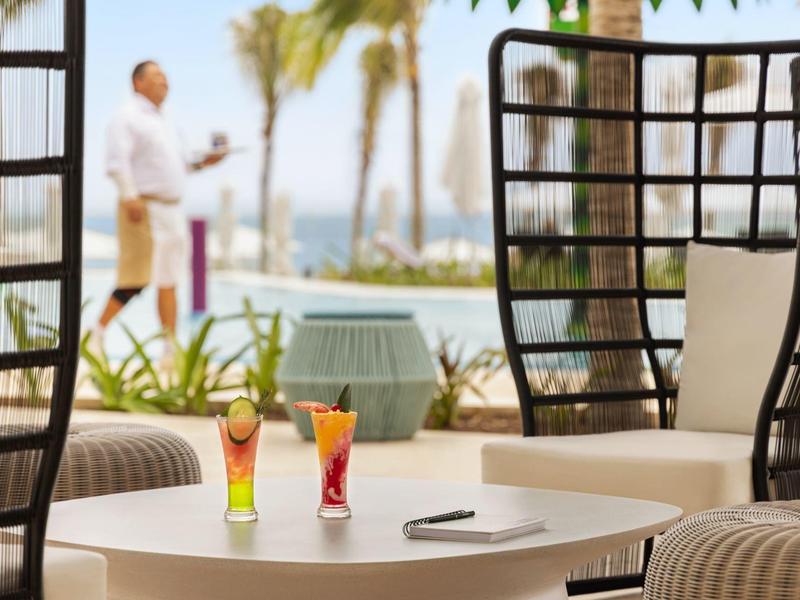 Zwei bunte Cocktails auf weißem Tisch, dahinter helle Sitzkörbe und Palmen am Strand.