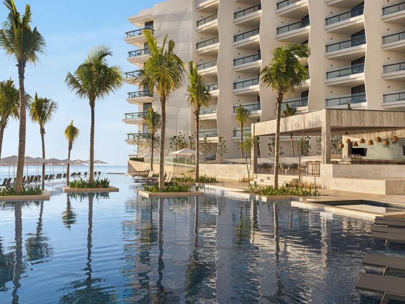 Moderner Hotelpool mit Palmen, Liegestühlen und Glasbalustraden bei klarem Himmel.