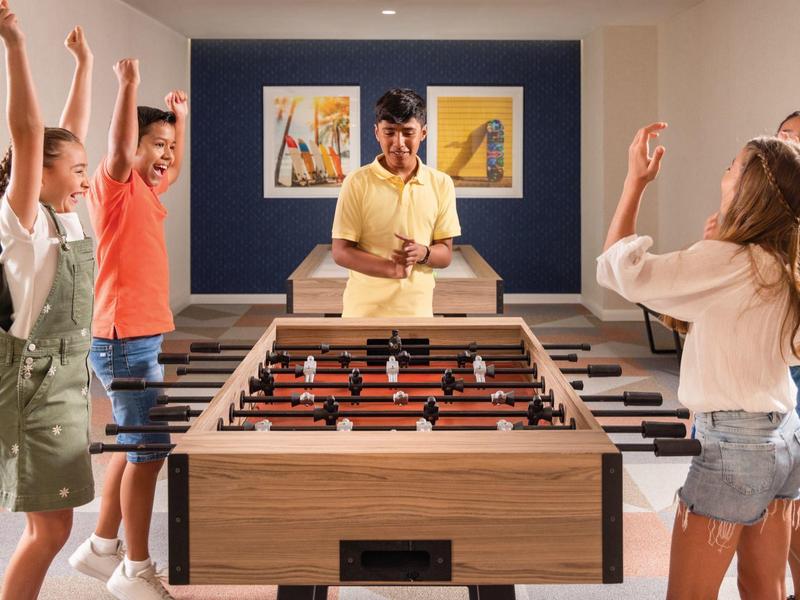 Fünf junge Menschen spielen und jubeln lebhaft um einen Kicker in einem modernen Raum.