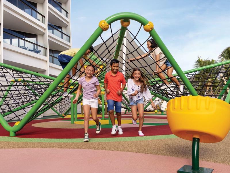Drei Kinder spielen vor einem grünen Klettergerüst auf einem bunten Spielplatz bei Wohnhäusern.