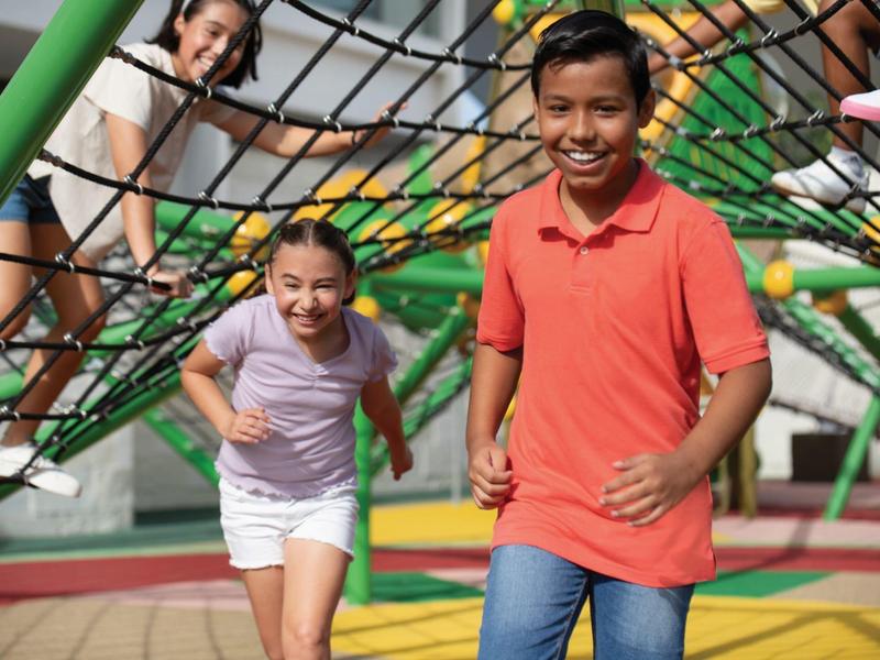 Drei lachende Kinder spielen im Klettergerüst auf einem farbenfrohen Spielplatz.