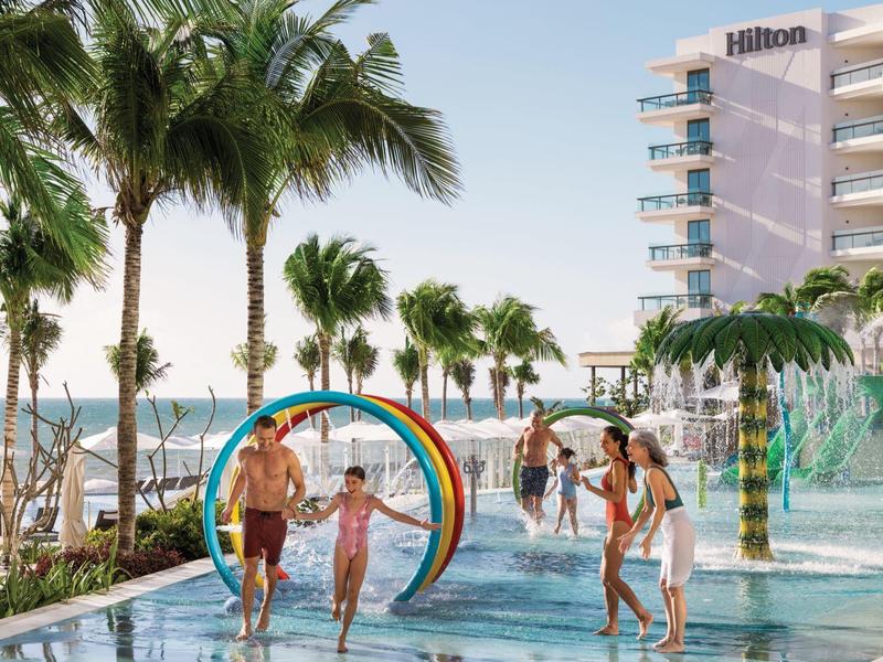 Familie spielt im flachen Pool mit Regenbogen-Wasserspiel, Palmen und Hotel im Hintergrund.
