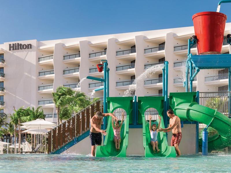 Kinder rutschen auf grüner Wasserrutsche in flachem Pool vor mehrstöckigem Hilton-Hotel.