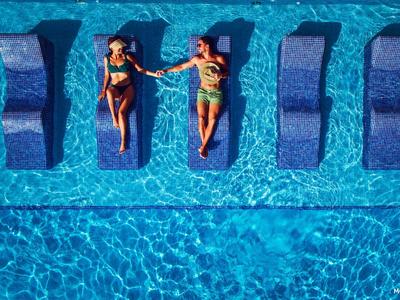 Dwie kobiety relaksują się na leżakach w czystym, niebieskim basenie, trzymając się za ręce.