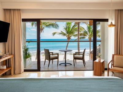 Blick aus einem Zimmer mit großem Fenster auf Balkon mit Tisch, Stühlen, Palmen und Meer im Hintergrund.