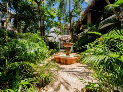 Zahrada s bujnými tropickými rostlinami a centrální fontánou mezi budovami.