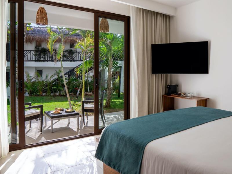 Světly hotelový pokoj s výhledem na zahradu, velkou postelí a televizí na stěně.