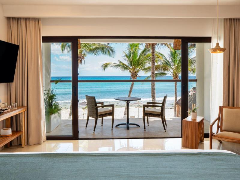 Hotelový pokoj s výhledem na moře, balkon se stolem a dvěma židlemi, palmy a pláž v pozadí.
