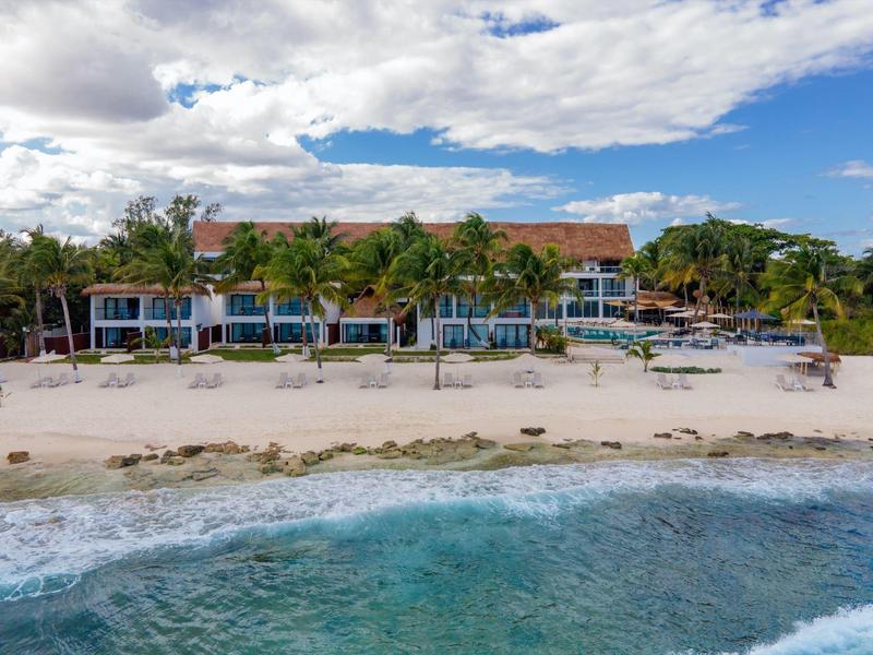 Hotelová budova s červenými střechami na bílé písečné pláži a palmami pod zataženou oblohou.