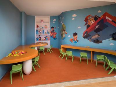 Sala giochi colorata per bambini con aree gioco, sedie verdi e murales di personaggi dei cartoni.