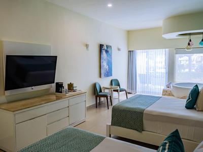 Camera d'hotel luminosa con due letti, TV, zona soggiorno e grandi finestre con tende.