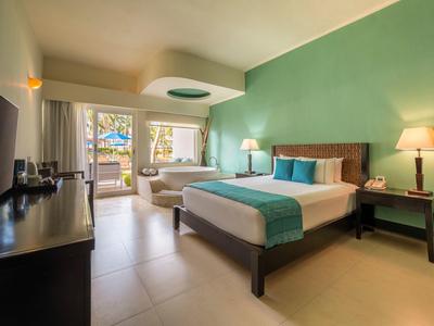 Camera d'hotel luminosa con letto matrimoniale, divano, lampade e accesso a una terrazza con vista mare.