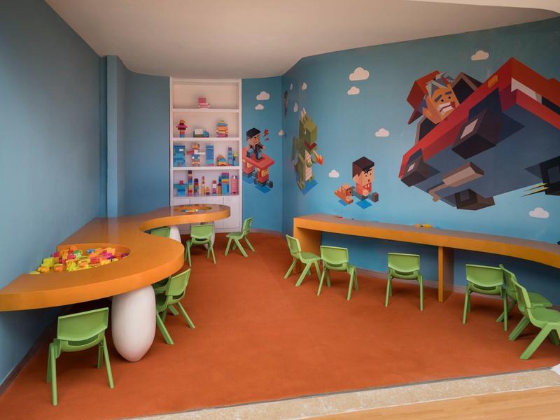 Sala giochi colorata per bambini con aree gioco, sedie verdi e murales di personaggi dei cartoni.