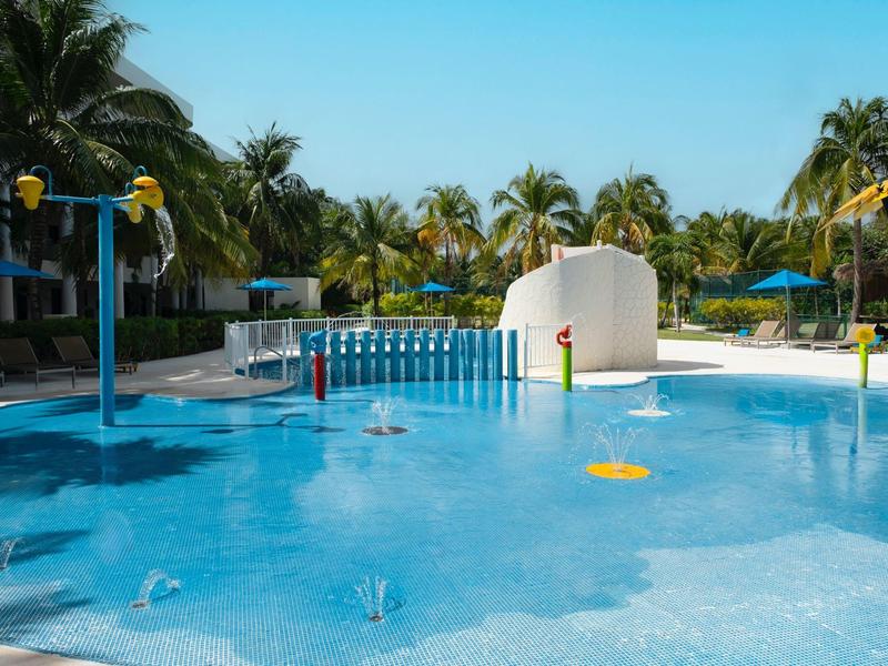 Piscina per bambini con giochi d'acqua e palme sullo sfondo in una giornata di sole.