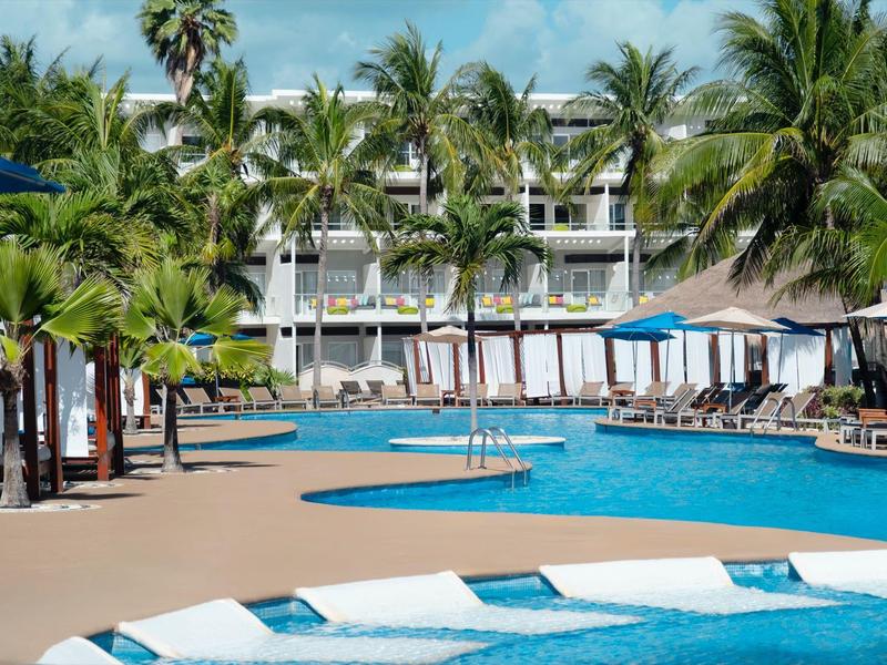 Piscina dell'hotel con lettini, ombrelloni e palme sotto un cielo azzurro.