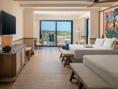 Camera d'hotel luminosa con due letti, TV, armadio e balcone con vista sul mare.