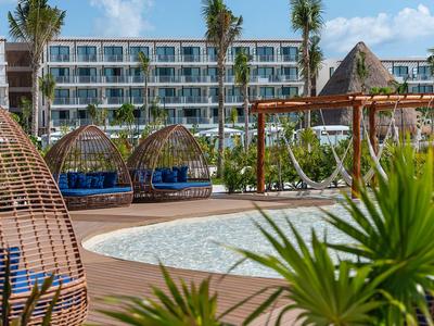 Modern hotel pool area with hammocks, wicker chairs, and surrounded by palm trees.