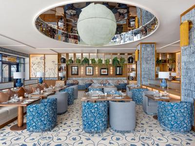 Modern hotel restaurant with blue upholstered poufs, wooden tables, and large ceiling mirror.
