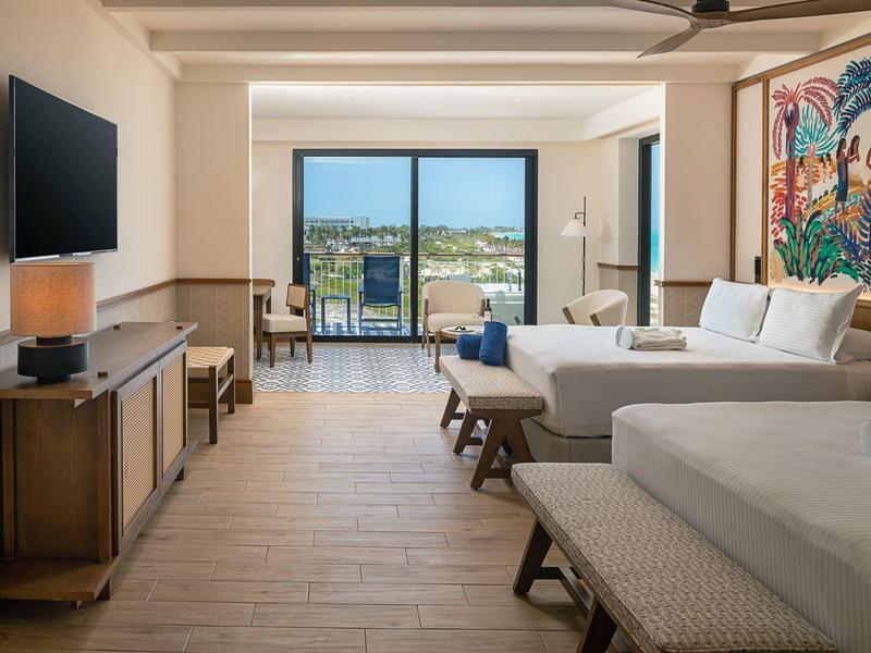 Camera d'hotel luminosa con due letti, TV, armadio e balcone con vista sul mare.