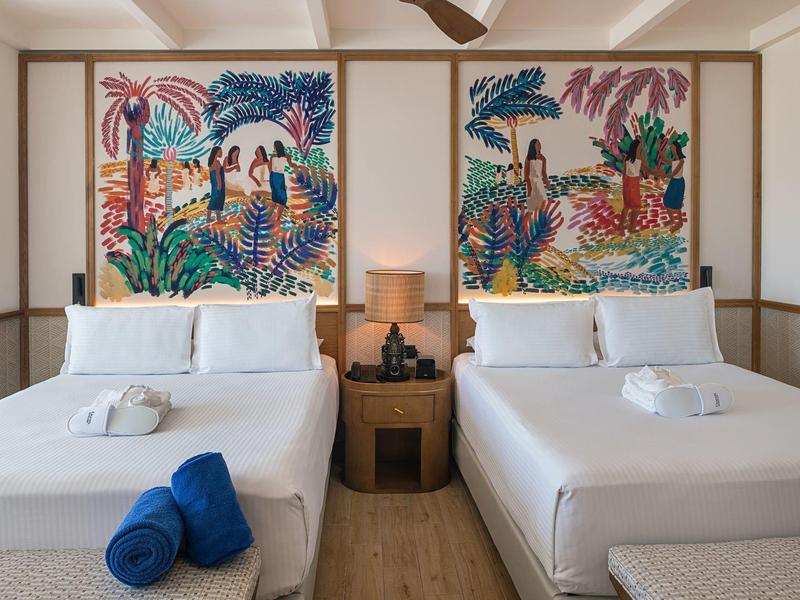 Camera d'hotel con due letti matrimoniali bianchi, arte murale colorata e un comodino al centro.