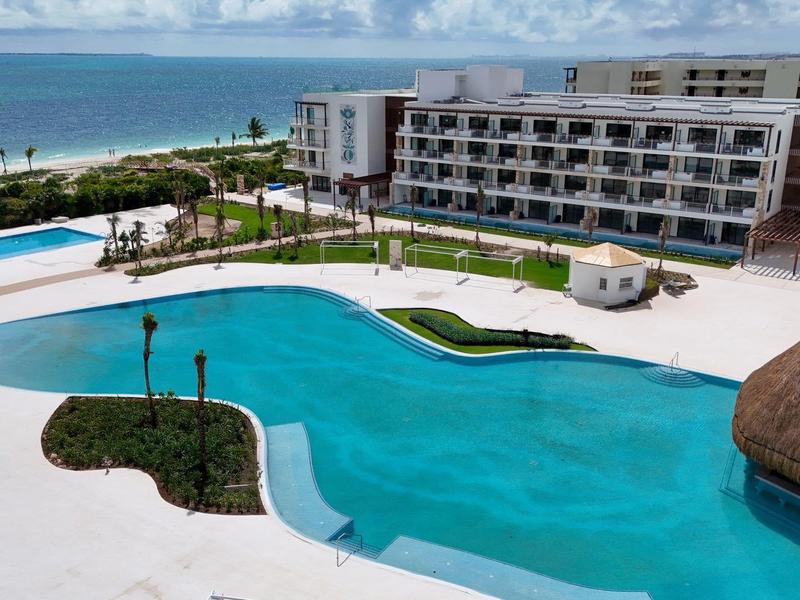 Complesso alberghiero moderno con grandi piscine vicino alla spiaggia e palme sotto un cielo azzurro.