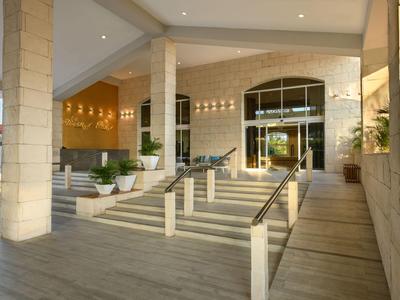 Moderne hotelentree met grote zuilen, trappen en brede deuren in warm licht.