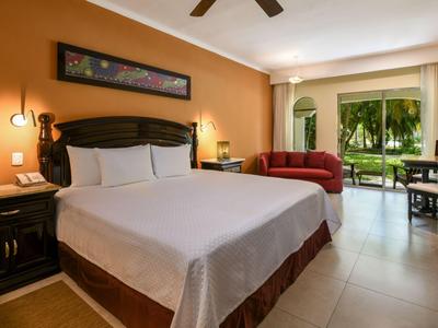 Pohodlný hotelový pokoj s manželskou postelí, nočními stolky, červenou pohovkou a výhledem na zelenou zahradu.