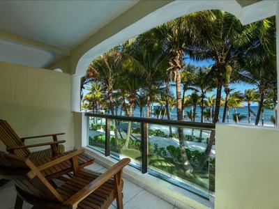 Balkon s dřevěnými židlemi a skleněným zábradlím s výhledem na palmy a moře.