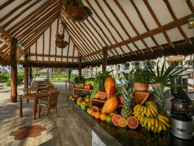 Krytý tržní stánek s čerstvým ovocem vedle otevřeného posezení v tropickém prostředí.