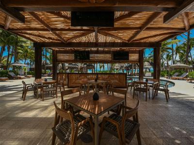 Padiglione aperto con soffitto in legno, tavoli e sedie, circondato da palme e spiaggia.