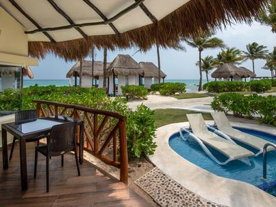 Vista da una terrazza con tavolo e sedie su una piscina, capanne sulla spiaggia e palme vicino al mare.