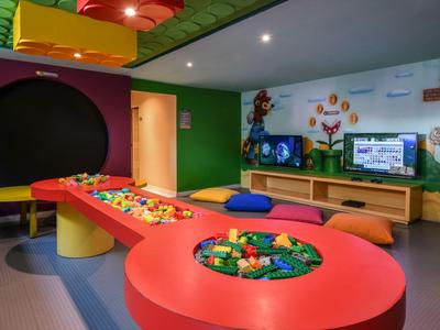Colorata sala giochi per bambini con tavoli, mattoncini LEGO e decorazioni murali in un hotel.