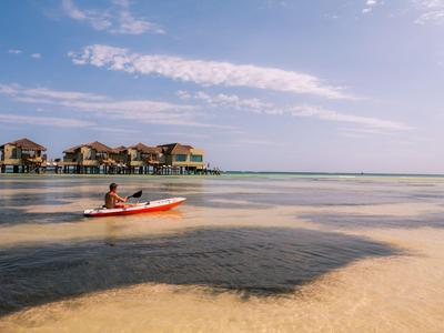 Osoba kajakem před vodními bungalovy za slunečného dne.
