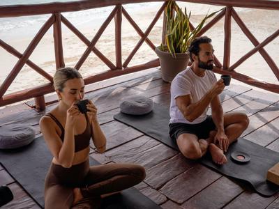 Dva lidé cvičí jógu na podložkách pod doškovou střechou u moře.