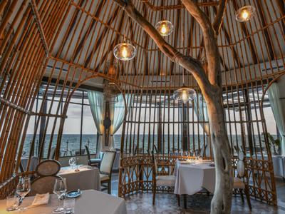 Restaurace s kulatou bambusovou konstrukcí, dřevěnou podlahou, bílými prostřenými stoly a přírodní atmosférou.