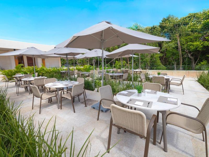 Area esterna moderna dell'hotel con tavoli, sedie e ombrelloni sotto un cielo azzurro.