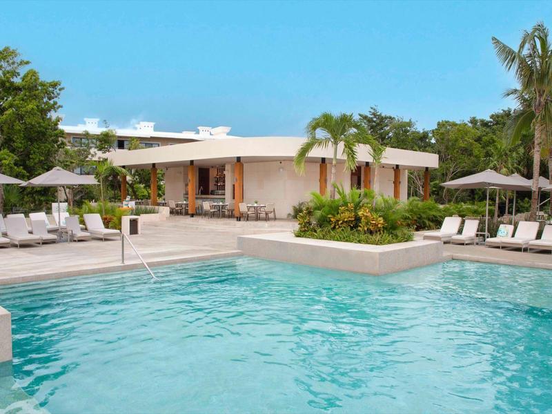 Piscina moderna dell'hotel con lettini, ombrelloni e palme tropicali sotto un cielo sereno.