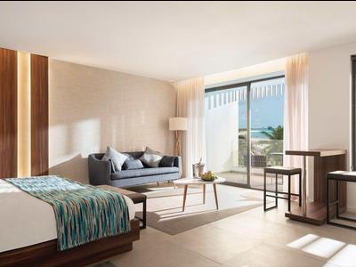 Moderní hotelový pokoj s velkými okny, posezením a postelí s modrými akcenty.