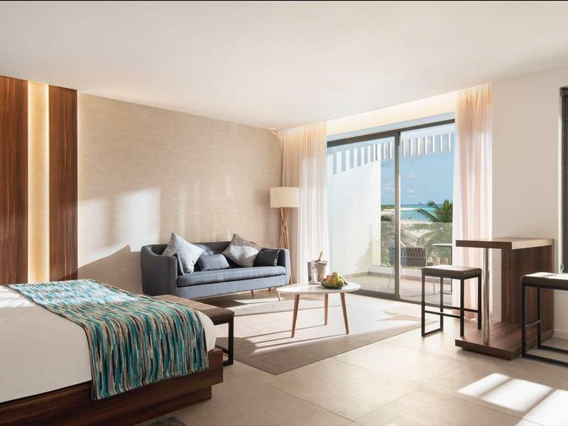 Moderní hotelový pokoj s velkými okny, posezením a postelí s modrými akcenty.