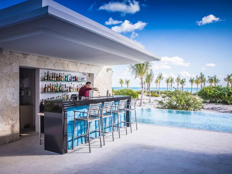 Moderní bar u bazénu s modrými barovými židličkami a výhledem na bazén a palmy.