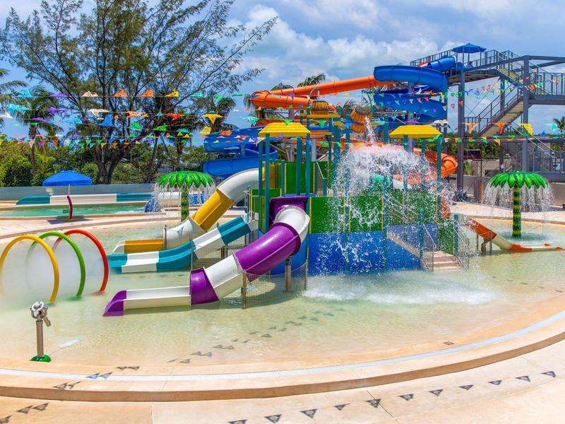 Parco acquatico colorato con scivoli, giochi d'acqua e piscina bassa sotto un cielo blu.