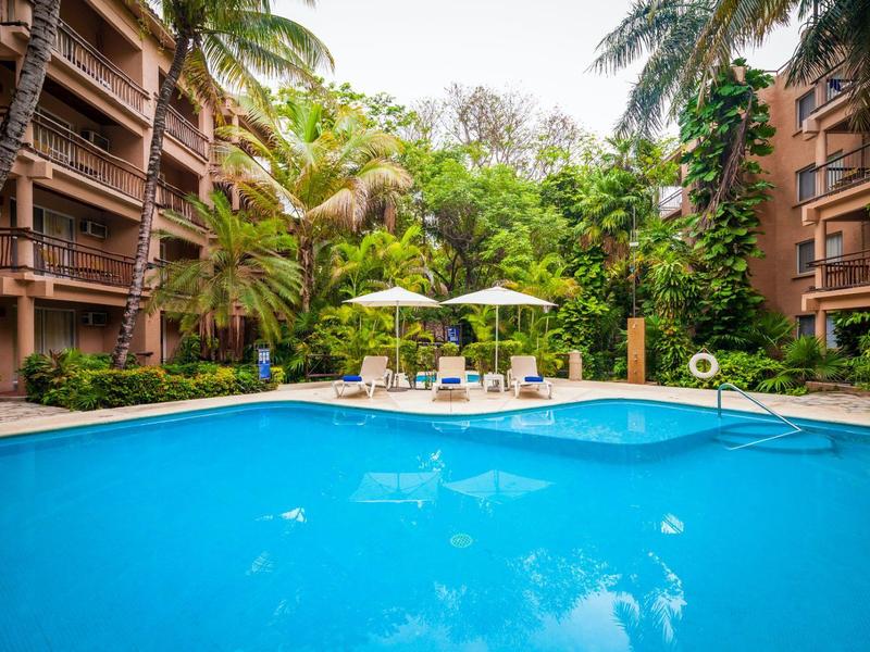 Piscina blu chiaro tra piante tropicali e hotel con lettini e ombrelloni.