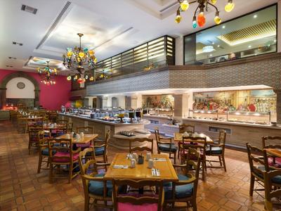 Grand restaurant avec tables en bois, chaises colorées et un grand buffet sous une galerie.