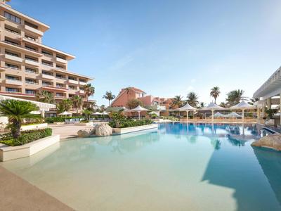 Großer Außenpool mit klarem Wasser, umgeben von Hotelgebäuden und Palmen unter blauem Himmel.